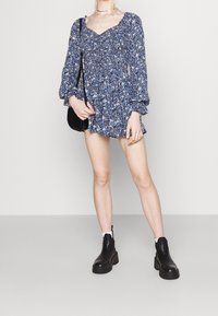 Květinové krátké šaty v modré barvě s dlouhými balónovými rukávy a čtvercovým výstřihem. Modelka má na sobě černé robustní boty a drží černou tašku.