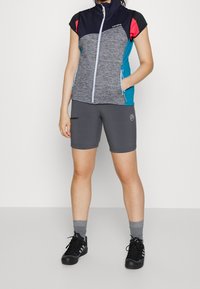 Sportig outfit bestående av en kortärmad, zip-försedd väst i grått, marinblått och teal, tillsammans med gråa tajta shorts och svarta skor.