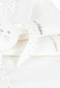Tissu blanc doux avec une tête de lapin ; présente des taches grises sur les oreilles, des accents brodés en vert et marron. Design simple et tactile.
