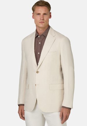 Blazer in lino beige chiaro con una vestibilità su misura, chiusura a un bottone e due tasche frontali. Indossato sopra una camicia marrone scuro.