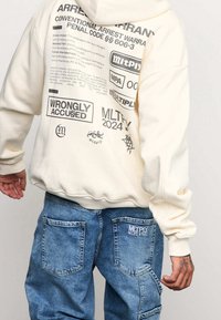 Krémová mikina s grafikou na zádech; doplněná světle modrými denimovými cargo kalhotami s kapsami na patche a logem na zadní straně.