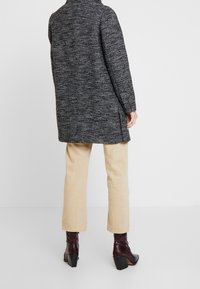 Lungo cappotto intrecciato grigio scuro con un motivo testurizzato, fessure laterali e colletto alto, abbinato a pantaloni beige chiaro e stivaletti alla caviglia bordeaux lucidi.
