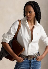 Femme aux cheveux tressés portant une chemise blanche boutonnée, un jean bleu, une ceinture décorative et un sac en cuir marron sur l'épaule.