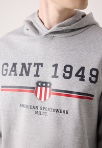 Hoodie gris avec un graphique sur le devant présentant "GANT 1949" en rayures noires et rouges. Comprend un emblème de bouclier avec trois étoiles et "AMERICAN SPORTSWEAR."