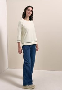 Helles Cremeoberteil mit Dreiviertelärmeln und schwarzem Saum, kombiniert mit blauen, ausladenden Jeans und hellen Sneakers.