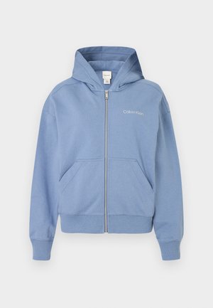 Mėlynas zip-up džemperis, pagamintas iš minkštos medžiagos, su gobtuvu, priekinėmis kišenėmis ir siuvinėtu logotipu. Standartinio kirpimo, su gumbai keliais ir apačioje.