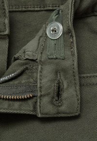 Detalj maslinasto zelene tkanine sa prozirnim dugmetom, rajsferšlusom i šivenim naglasima. Sadrži petlju i mali pocepan deo. Teksturirana površina.