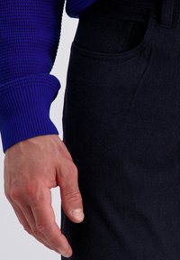 Blauwe textuur trui mouwen en donkere stoffen broek worden getoond, met een hand die tegen de zak van de broek rust.