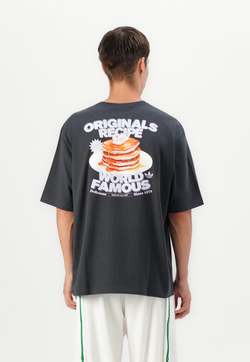 Sort oversized t-shirt med et farverigt grafisk motiv af pandekager på ryggen med teksten "ORIGINALS RECIPE WORLD FAMOUS." Korte ærmer.