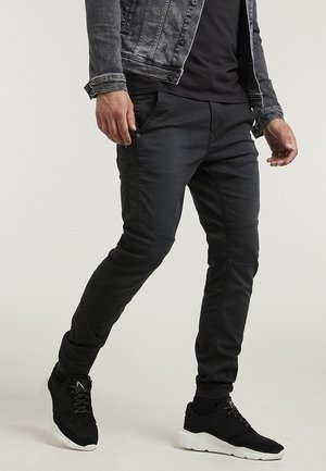 Mann trägt schwarze schmal geschnittene Hose, schwarze Sneaker mit weißen Sohlen, schwarzes Hemd und dunkelgraue Denimjacke, steht mit einem Bein nach vorne.