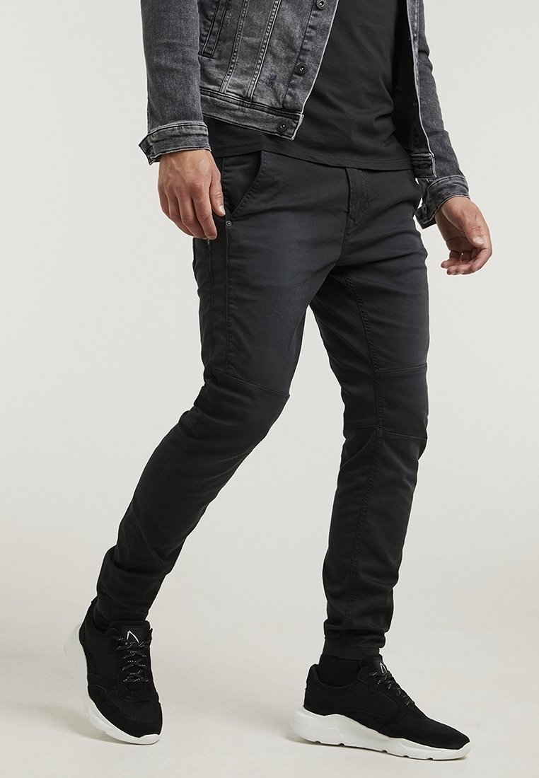 Homme portant un pantalon noir ajusté, des baskets noires à semelles blanches, une chemise noire et une veste en jean gris foncé, debout avec une jambe avancée.