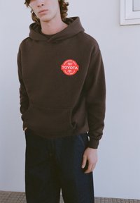Hnedá mikina s kapucňou a predným vreckom, s červeným kruhovým logom Toyota. Nosená s tmavomodrými rifľami. Hladká textúra látky.