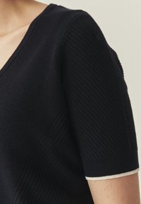 Top negro de punto con escote en V y mangas cortas, que presenta un patrón acanalado texturizado y un detalle blanco en el dobladillo de la manga.