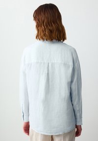 Himmelblaues Leinenhemd mit langen Ärmeln, Rückenfalten, und geknöpften Manschetten, das einen abgerundeten Saum und eine glatte Textur aufweist.