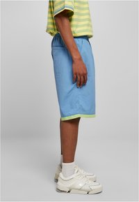 Gaiši zili šorti ar zaļu malu, izgatavoti no teksturēta auduma. Piedāvā sānu kabatu un elastīgu jostasvietu. Valkāti ar baltām sporta kurpēm.