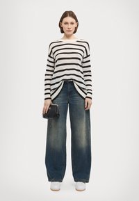 Pull rayé crème et noir, associé à un jean large bleu, un petit sac à main noir et des baskets blanches. Matériaux texturés, coupe décontractée.