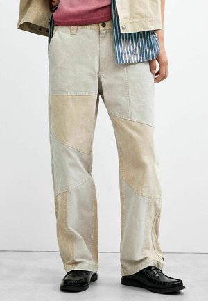 Beige patchworkbroek gedragen door een persoon, gecombineerd met zwarte loafers op een lichtgrijze vloer en een witte achtergrond.