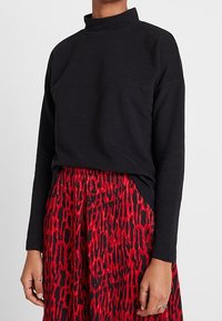 Col roulé côtelé noir à manches longues, associé à une jupe rouge mi-longue avec un motif abstrait noir.