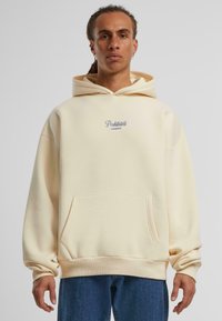 Šviesiai smėlinis hoodie su priekyje esančia kišene, raukšlėtomis rankovėmis, tamsiai mėlynu siuvinėtu logotipu "Prohibited Garments", minkšta tekstūra ir laisvas fasonas.