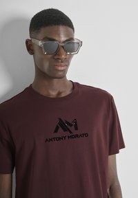Hombre que lleva gafas de sol de gran tamaño con montura transparente y una camiseta color burdeos con el logo negro "Antony Morato" sobre un fondo blanco.