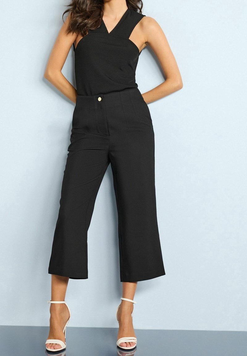 Combinaison sans manches noire avec un design croisé, taille haute et jambes larges et raccourcies. Comprend un bouton à l'avant et une texture de tissu lisse.