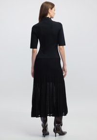 Robe noire avec un haut ajusté à manches courtes et une jupe évasée plissée. Portée avec des bottes marron brillantes jusqu'aux genoux.