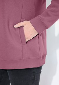 Felpa a maniche lunghe di colore viola chiaro con polsini e orlo a coste, dotata di una tasca laterale sulla sinistra. Tessuto morbido con texture liscia.