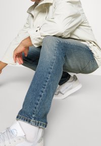 Giacca beige chiaro sopra jeans blu, con taglio dritto e texture sbiadita, abbinata a scarpe da ginnastica bianche e calze bianche.