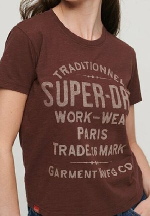 Osoba w brązowej koszulce Super-Dry z wyblakłym białym napisem „Traditionnel Super-Dry Work-Wear Paris Trade Mark Garment Mfg.Co".