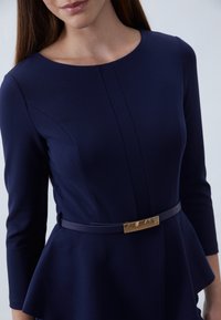 Granatowa sukienka peplum z długimi rękawami, okrągłym dekoltem, teksturowanym materiałem oraz cienkim granatowym paskiem z akcentem w postaci złotej prostokątnej klamry.