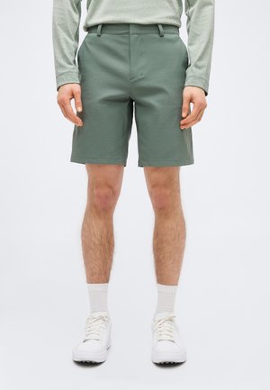 Hombre vistiendo pantalones cortos verdes a medida, una camiseta de manga larga, calcetines blancos y zapatillas blancas, de pie contra un fondo liso.