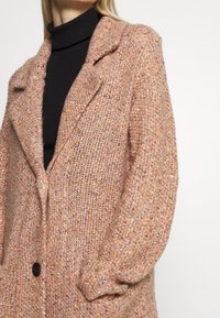 Abrigo de punto color durazno con un patrón texturizado, grandes solapas y un solo botón negro. Presenta mangas largas y relajadas.