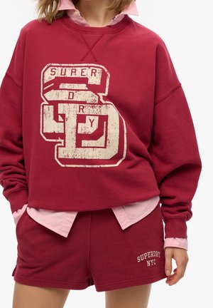 Osoba ubrana w czerwoną bluzę Superdry z dużym logo oraz dopasowane szorty z napisem "Superdry NYC", na jasnoróżowej koszuli.