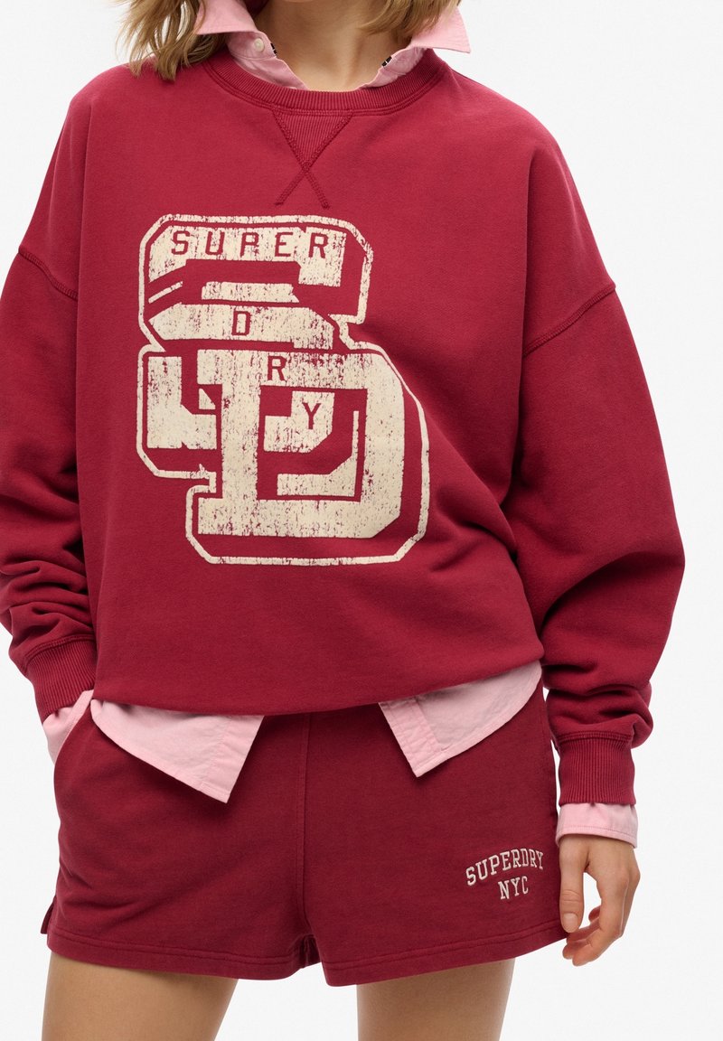 Personne portant un sweat-shirt rouge Superdry avec un grand logo et un short assorti avec le texte "Superdry NYC", superposé sur une chemise rose clair.
