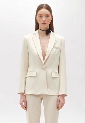 Blazer beige su misura con rever a punta, chiusura a un bottone e tasche frontali. Indossato con un choker nero con una rosa.