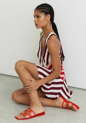 Moteris su pintinėmis plaukais sėdi sukryžiuotomis kojomis, vilkinti rankovių neturinčią dryžuotą suknelę ir raudonas atvirų pirštų plokščias basutes ant pilko grindinio.