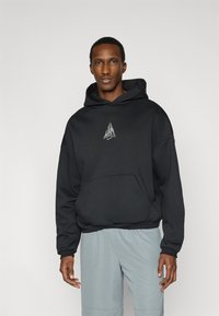 Μαύρο φούτερ με κουκούλα από μαλακό ύφασμα, με μπροστινή τσέπη και έμβλημα στο στήθος. Συνδυάζεται με ανοιχτό γκρι σορτς.