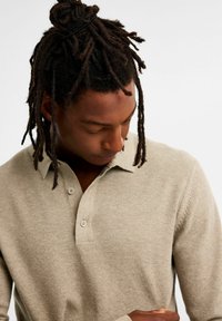 Beige, texturerad stickad polo med tre knappslåar, krage och ribbade ärmmuddar, bärs av en person som ser ner.