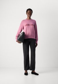 Donna che indossa un maglione rosa di Calvin Klein, jeans neri e ballerine nere, mentre tiene una grande borsa nera su uno sfondo bianco uniforme.