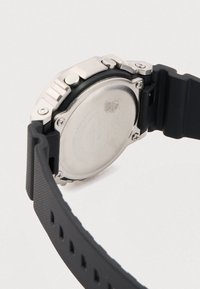 Μαύρο λουράκι ρολογιού από καουτσούκ με υφή στην επιφάνεια, μεταλλική θήκη και λείο ασημί πίσω μέρος που εμφανίζει χαραγμένες λεπτομέρειες και λογότυπα.