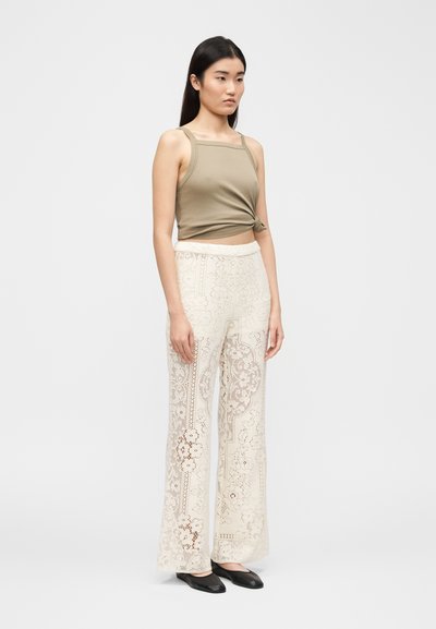 Γυναίκα με χακί αμάνικη μπλούζα με κόμπο μπροστά, κρεμ φαρδιά παντελόνια με δαντέλα και μαύρα επίπεδα παπούτσια, στέκεται μπροστά σε απλό φωτεινό φόντο.