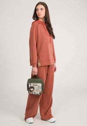 Mujer con sudadera y pantalones de color óxido sosteniendo una bolsa verde y beige con diseño de dibujos animados, de pie contra un fondo claro y liso.