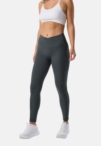 Kobieta ubrana w ciemnoszare legginsy z wysokim stanem, biały sportowy biustonosz i białe buty sportowe, stojąca na tle jasnego jednolitego tła.