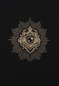 Un tejido negro presenta un bordado dorado central de un ojo rodeado de patrones elaborados y líneas radiantes, creando un diseño detallado.