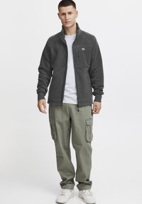 Gray fleece jacket with high collar, zippered chest pocket, paired with olive cargo pants and white sneakers. Smooth texture, casual fit.