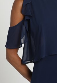 Marinblå klänning med kall axel-design och lager av volanger på ärmarna, tillverkad av ett lätt och genomskinligt tyg.