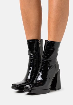 Zwarte enkelboots met hakken en een glanzende lakafwerking, voorzien van een krokodillenstructuur en een zijrits voor gemakkelijk aan- en uittrekken. Vierkante neusdesign.
