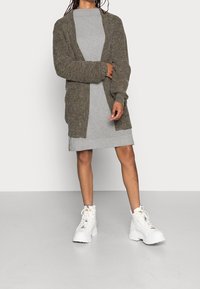 Pletený olivově zelený cardigan přes světle šedé žebrované šaty. Model má na sobě bílé robustní boty se žlutými tkaničkami. Jednoduchý design s otevřeným předním dílem.