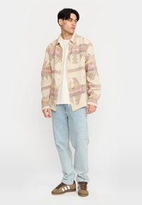 Beige gewebtes Hemd mit pinken und braunen geometrischen Mustern, kombiniert mit hellblauen Jeans und braunen Sneakers mit weißen Streifen.