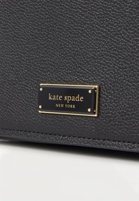 Tekstuuriga must nahk koos kuldse tooni brändi sildiga, millel on tekst "kate spade NEW YORK". Omab nähtavat õmblust ja struktureeritud kuju.